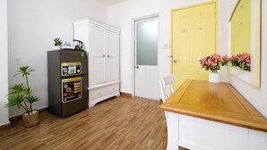 Quarto com piso de madeira, paredes brancas, guarda-roupa branco, porta amarela, uma pequena mesa com um vaso de flores e uma geladeira com visor digital.
