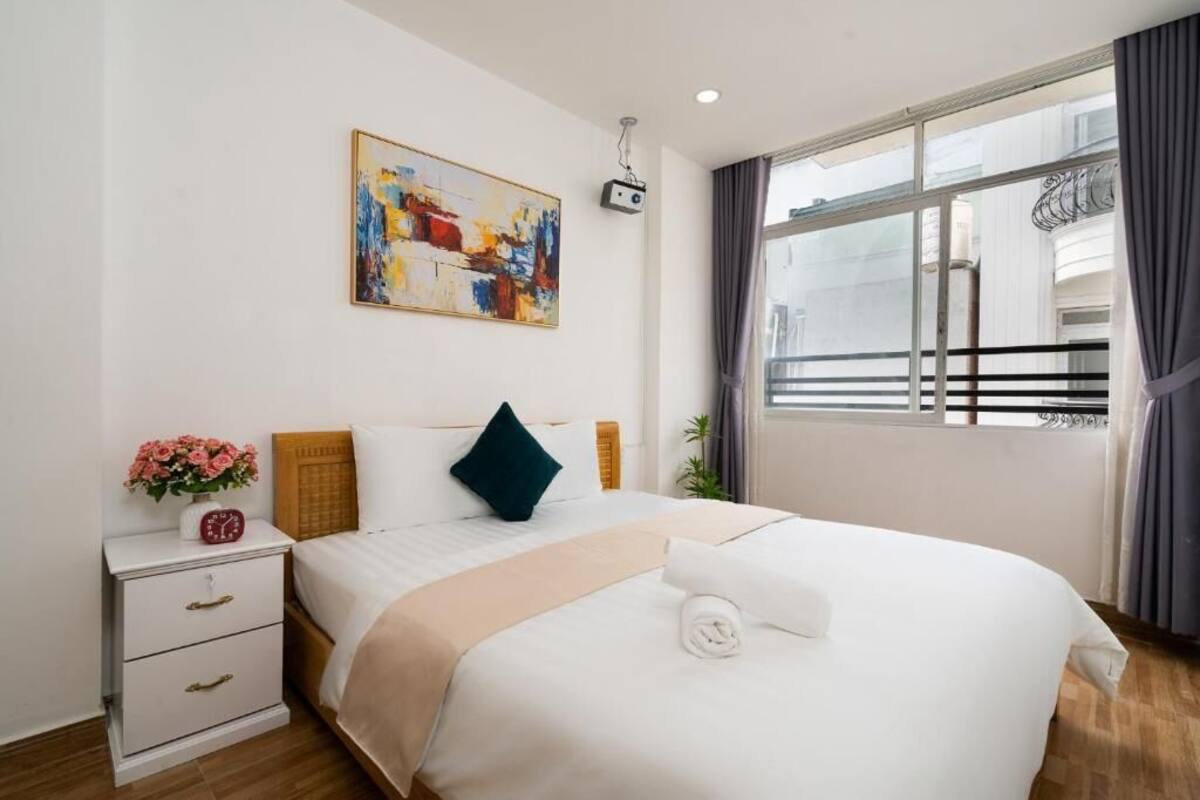 Een hotelkamer met een bed, nachtkastje, schilderij, raam met gordijnen en uitzicht op het exterieur.