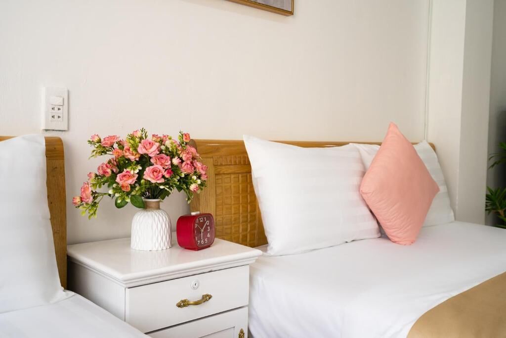 A bedroom with a bed, a nightstand, a vase of flowers, a clock, and a power outlet.