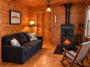 A cozy cabin interior with a sofa, a rocking chair, a wood-burning stove, and a framed fish illustration on the wall. - Hambrook Point Cottage | Renous & Miramichi Rivers (Renous)