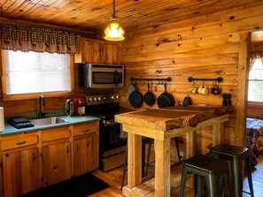 A wooden cabin kitchen with a microwave, stove, and hanging pots. - Hambrook Point Cottage | Renous & Miramichi Rivers (Renous)