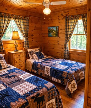 A cabin-style room with two beds, plaid curtains, a ceiling fan, a painting, and a window with a view of trees. - Hambrook Point Cottage | Renous & Miramichi Rivers (Renous)