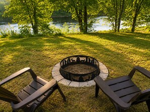 A circular fire pit with a metal design, surrounded by a stone border, with two wooden Adirondack chairs facing it.
