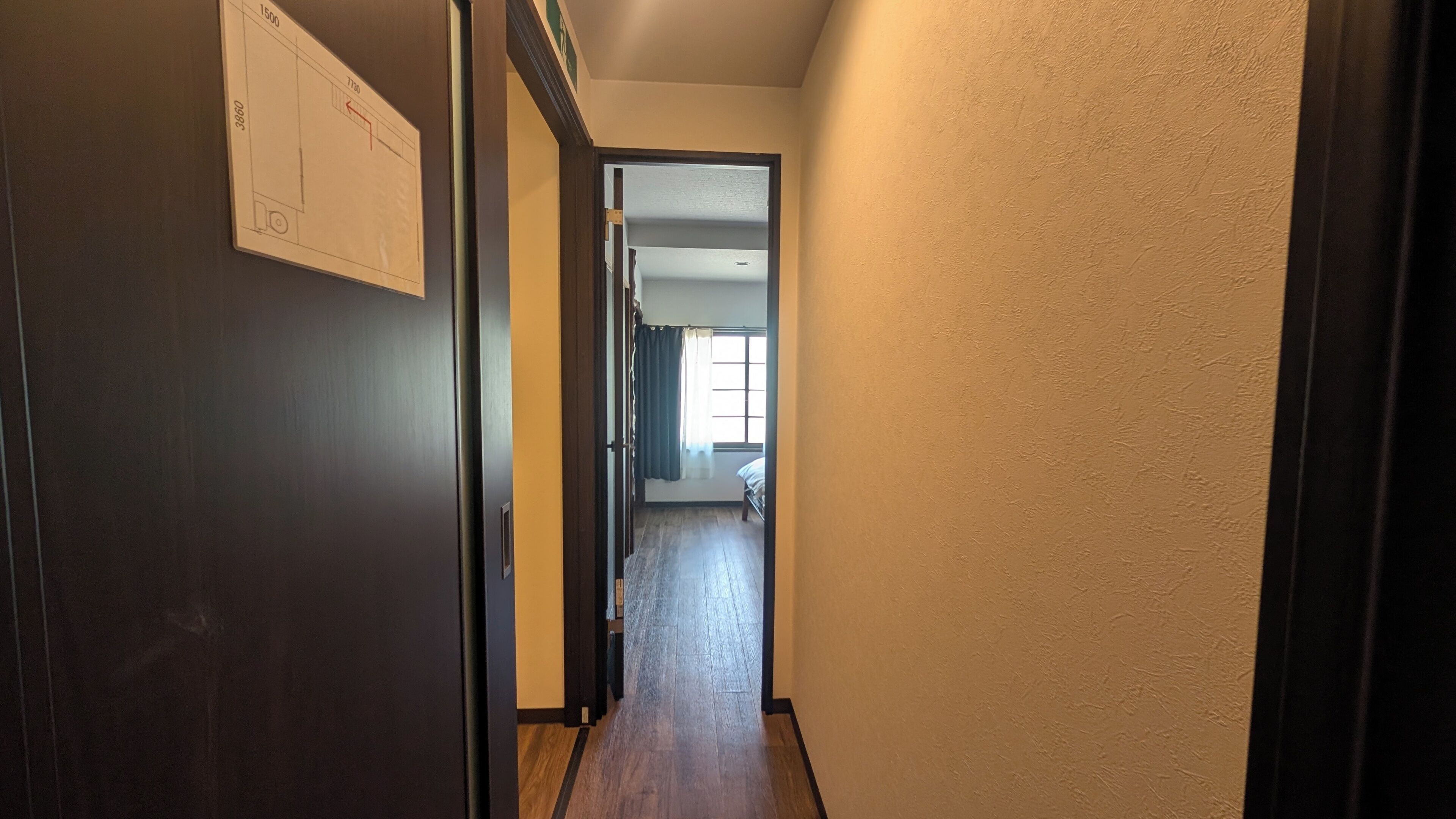A hallway with a door on the left, a window with curtains, and a bed in the room at the end.