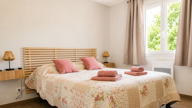 Un dormitorio con cabecera de madera, dos lámparas de noche, colcha con estampado floral y una ventana con cortinas.