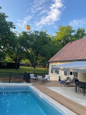 Ein Schwimmbecken mit Holzterrasse, Liegestühlen und einem Haus mit einem Ziegeldach.