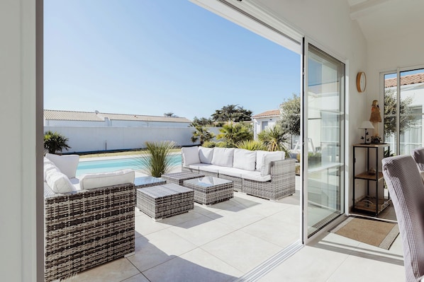 A balcony with wicker furniture, a white sofa, and a glass door leading to a pool. - Villa Tikaz 4* Ile d'Oléron 8 people swimming pool (Saint-Denis-d'Oléron)