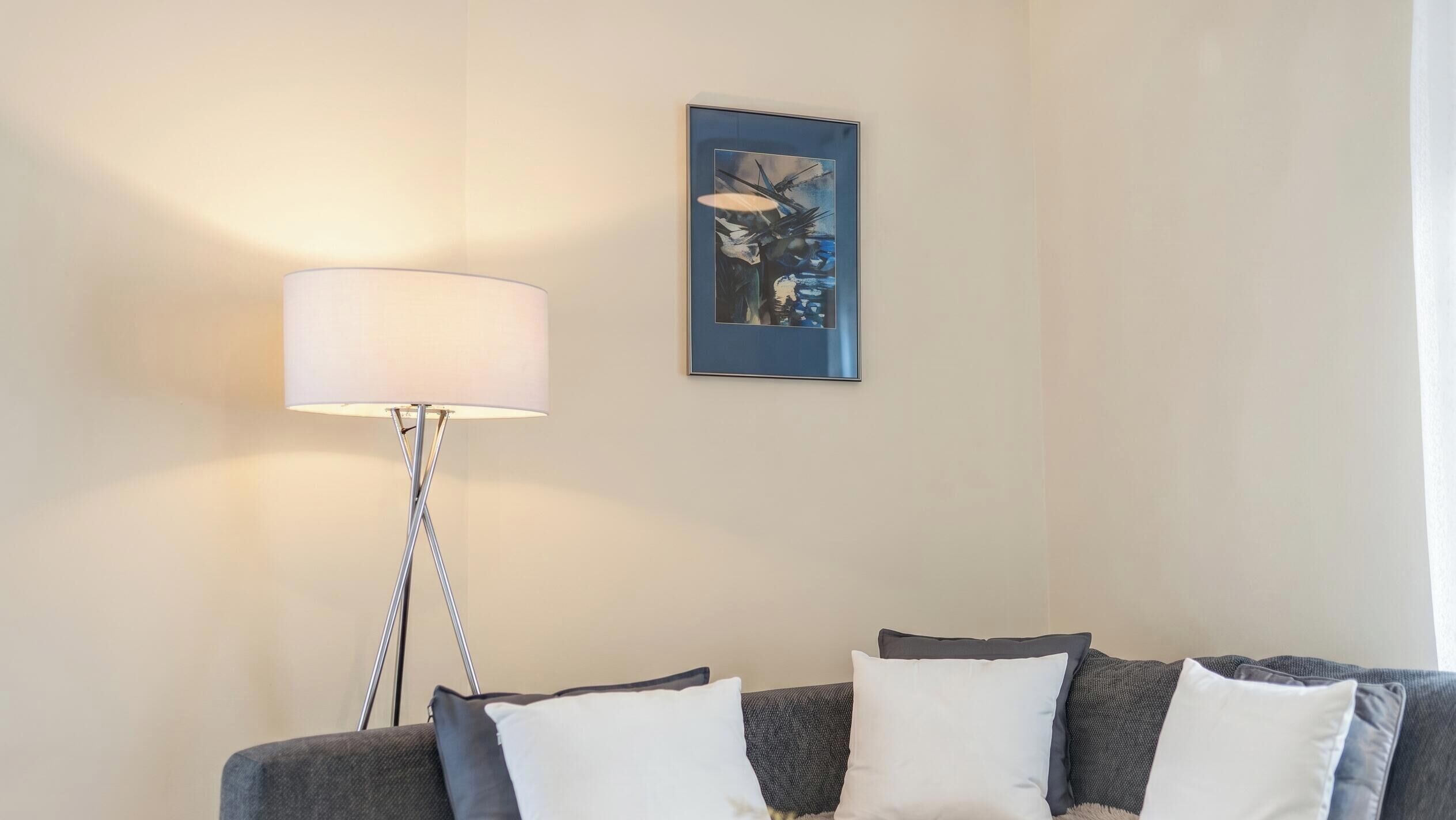 A modern living room with a grey sofa, white and grey cushions, a floor lamp, and a framed abstract painting on the wall.