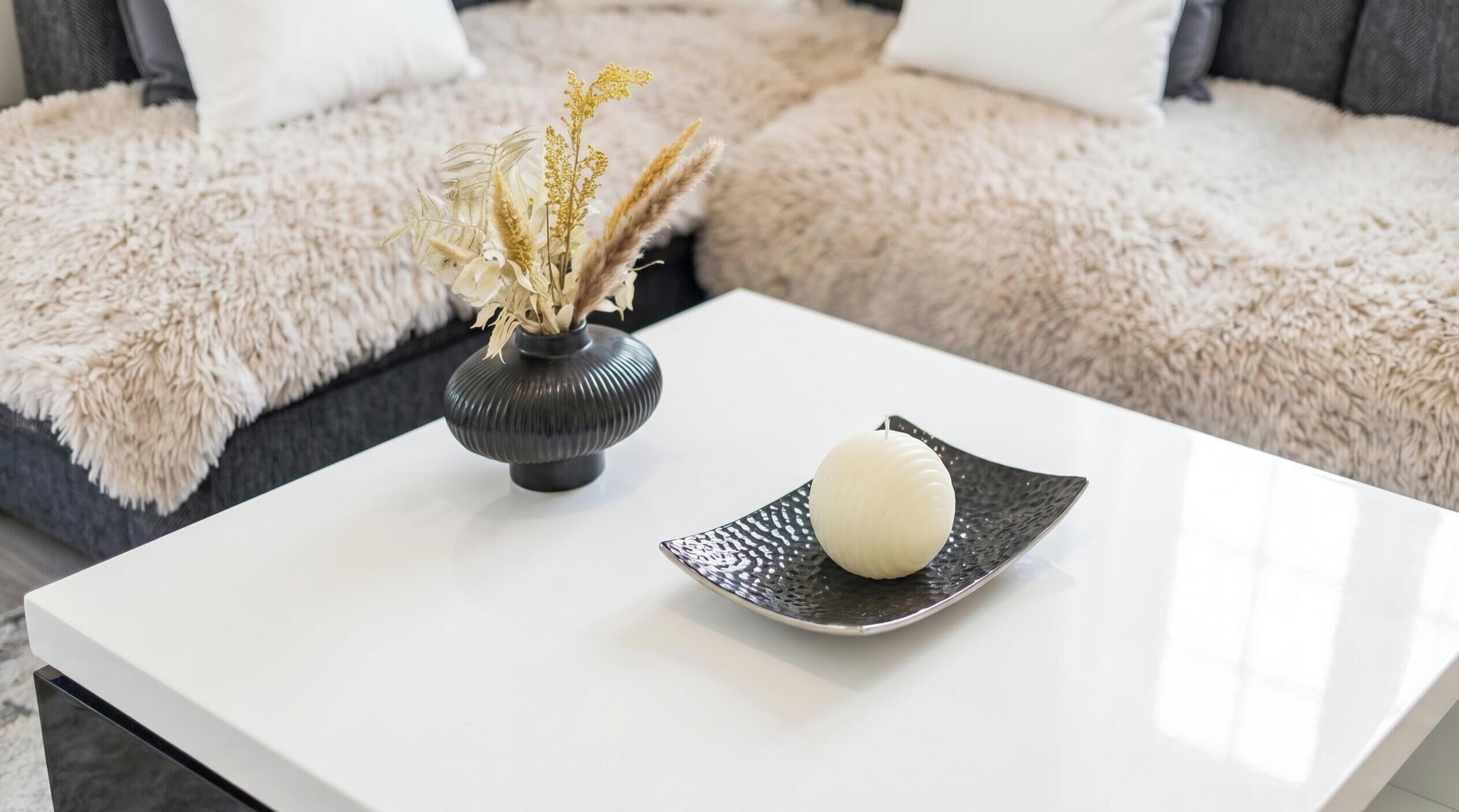 A white coffee table with a vase of dried flowers and a single candle on a textured plate.