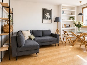 Ein modernes Wohnzimmer mit einem grauen Ecksofa, Holzboden und einem Bücherregal mit Büchern und Dekorationselementen.