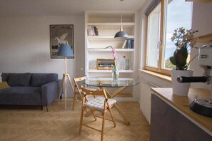 Ein modernes Wohnzimmer mit Glastisch, Holzstühlen, einer Couch, einem Bücherregal und einem Fenster mit Blick.