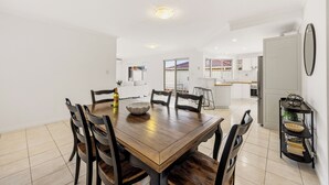 Un grand espace à manger avec une table et des chaises en bois, une cuisine à l’arrière-plan et une porte-fenêtre coulissante donnant sur une terrasse extérieure.