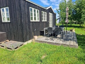A black wooden building with white-framed windows, a patio with a table and chairs, and a grassy yard. - 2 Person Holiday Home in Dannemare (Dannemare)