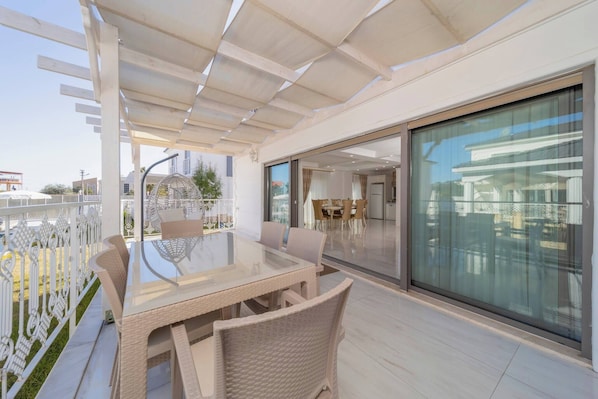 An outdoor patio with a glass table and wicker chairs, a pergola overhead, and a view of the cityscape through large windows.