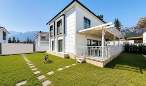 A modern two-story house with a covered patio, a well-maintained lawn, and a mountain backdrop.