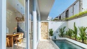 Un área de piscina moderna al aire libre con suelo de piedra, una mesa de comedor con sillas y plantas en macetas.