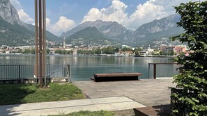 A lakeside promenade with a wooden bench, metal railings, and a view of a town and mountains. - Second Lake House - Affitti Brevi Italia (Malgrate)