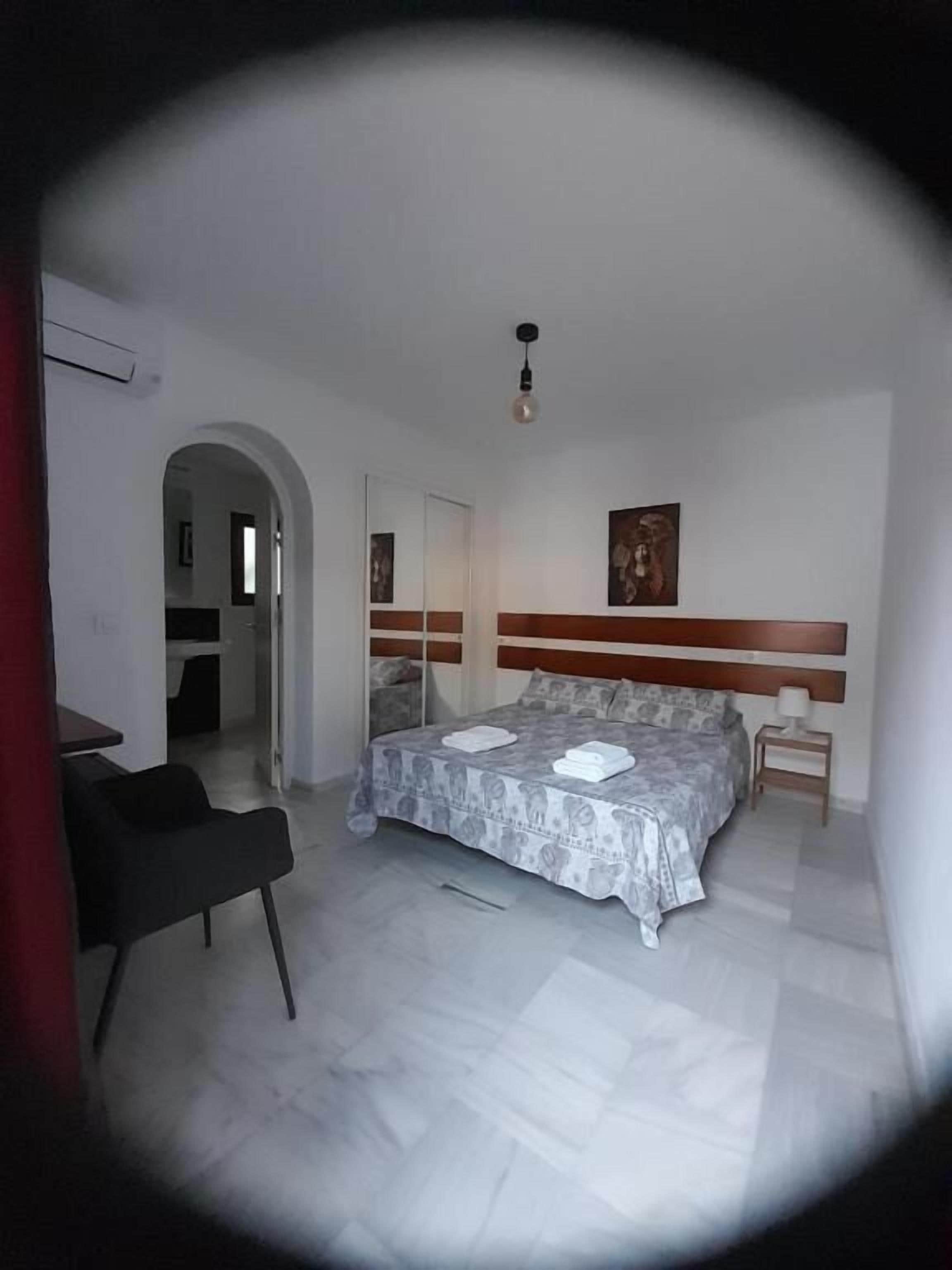 A bedroom with a bed, a chair, a bedside table, and a door leading to another room.