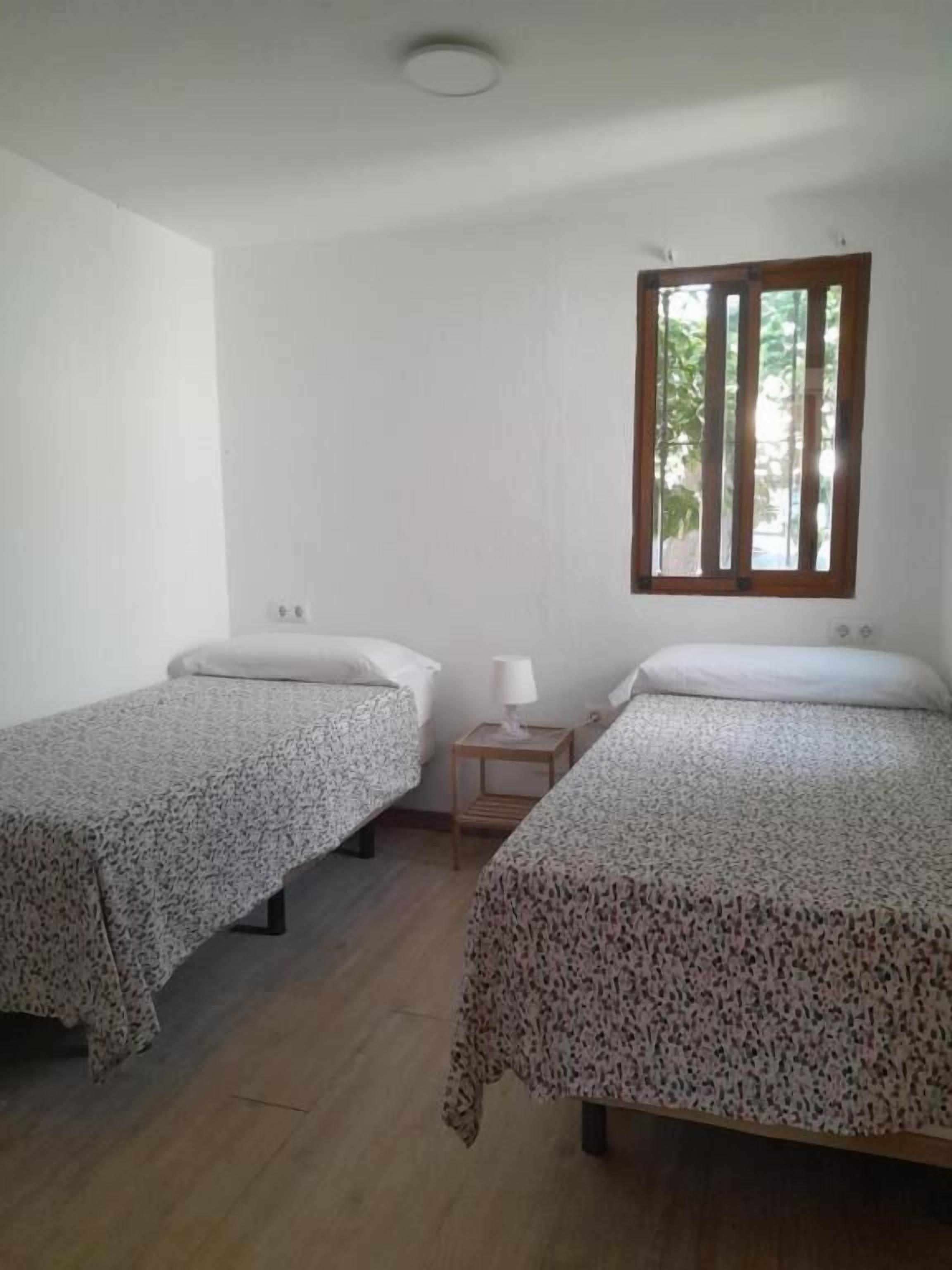 Una habitación con dos camas individuales, una mesita de noche pequeña con lámpara, un ventanal con marco de madera y una pared blanca.