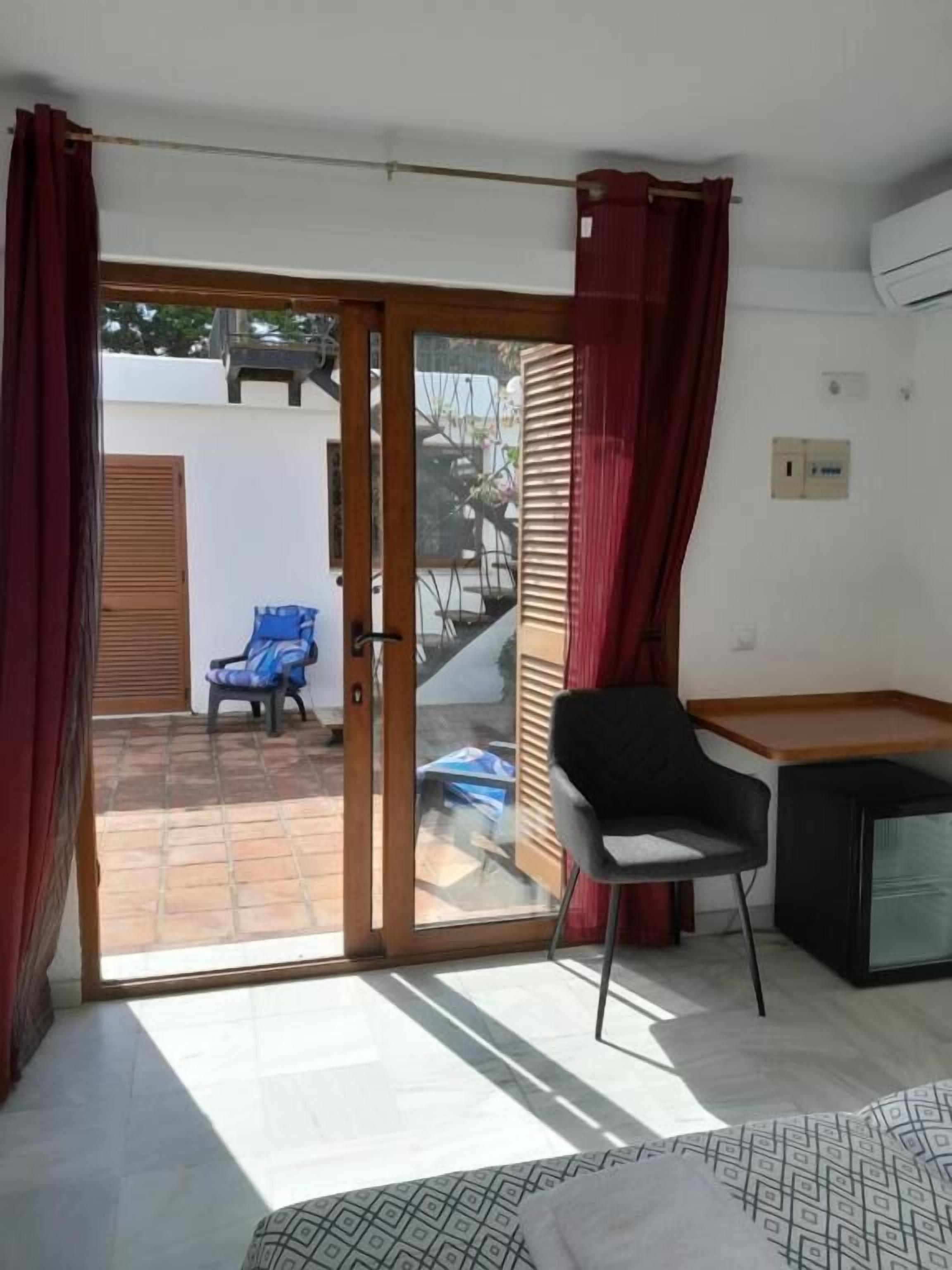 Una habitación con cama, escritorio, silla y puerta corrediza de vidrio que da a un espacio exterior con una silla y una mesa.