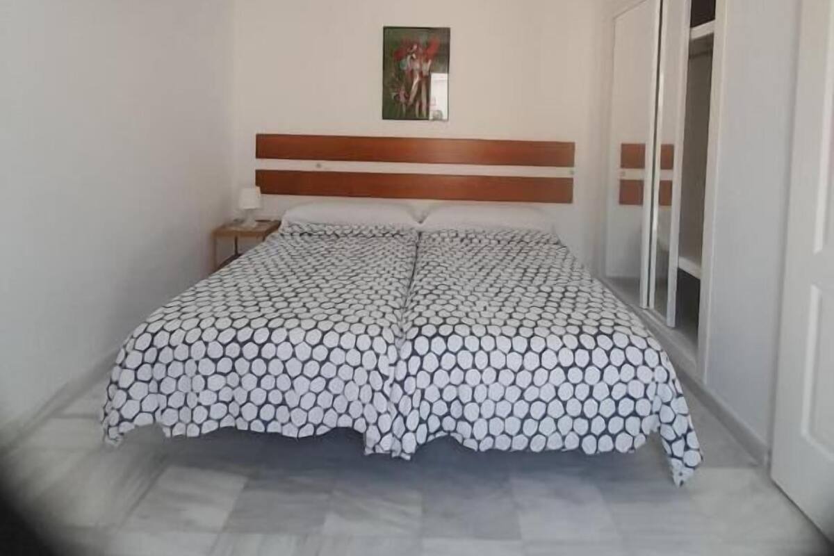 A bedroom with a bed, a headboard, a nightstand, a wall-mounted light, and a window.
