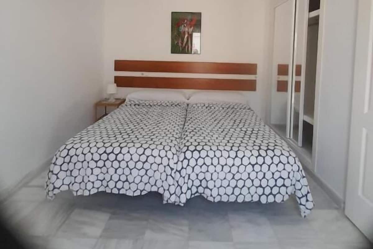 Une chambre à coucher comprenant un lit, une tête de lit, une table de chevet, une lampe fixée au mur et une fenêtre.