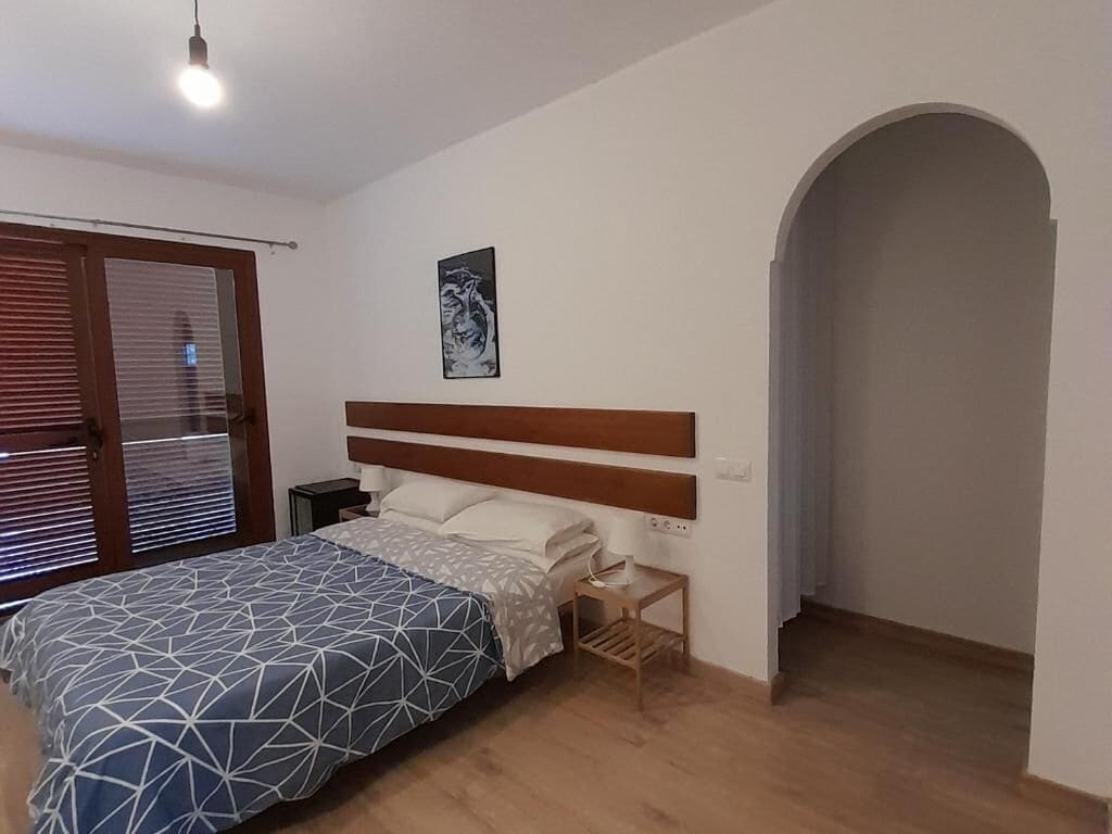 Un dormitorio con una cama, una mesita de noche, un cabecero de madera, un cuadro enmarcado en la pared y un umbral que da a otra habitación.