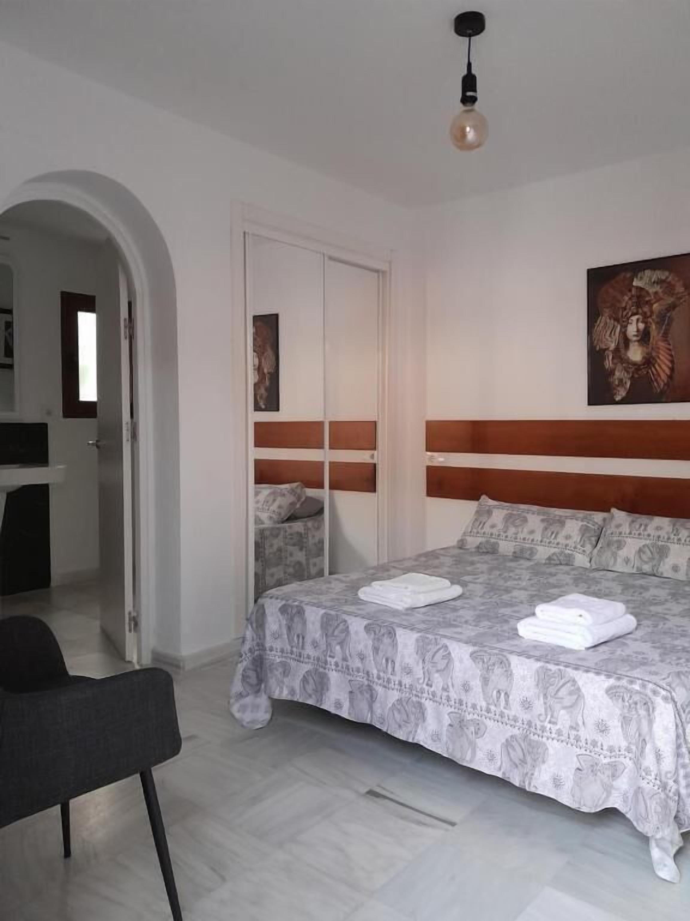 Un dormitori amb llit, cadira, armari i un quadre a la paret.