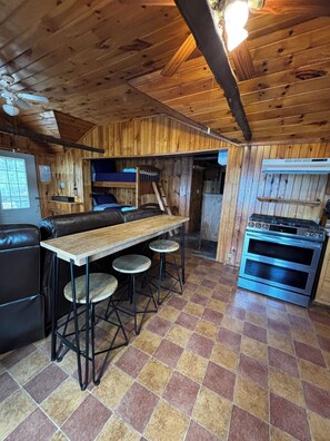 A wooden cabin interior with a kitchen area featuring a stove, a dining table with stools, and a bunk bed.