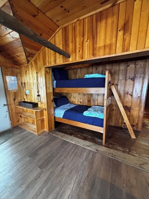 Una cama litera con cabecera y piecera de madera, un estante pequeño y suelo de madera.