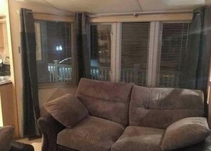 A corner room with a sofa, a window with blinds, and a partial view of a kitchen area. - Waterside k5 With Wraparound Deck Opposite Fishing Lake (Skegness)