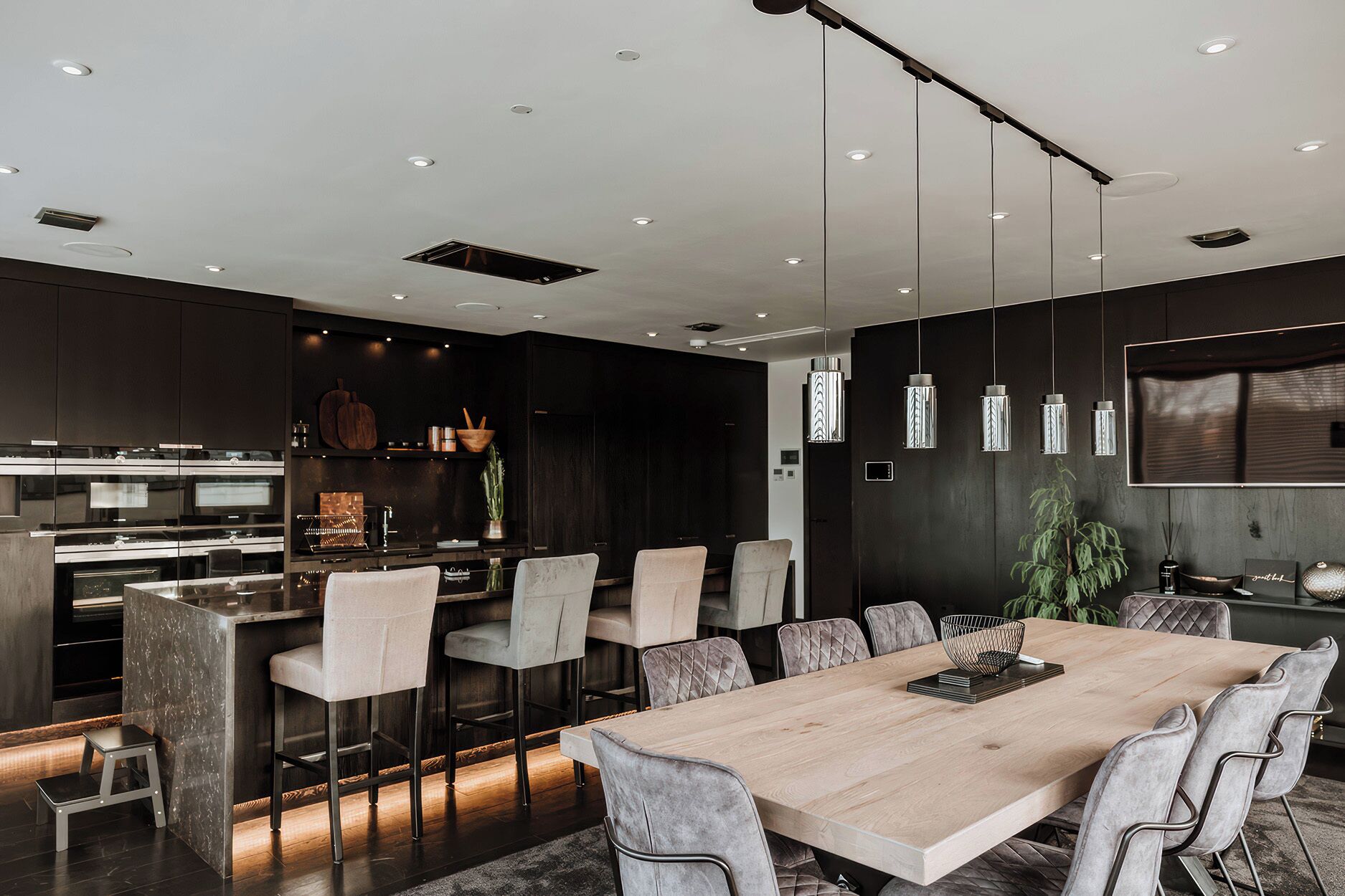 Une cuisine moderne dotée d’appareils électroménagers intégrés, un coin repas avec une grande table en bois et des chaises rembourrées, ainsi qu’un éclairage suspendu.