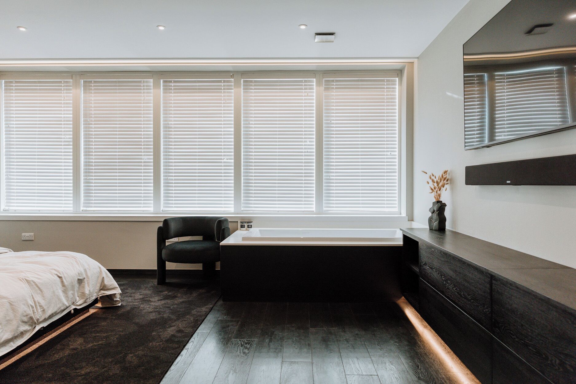 Une chambre moderne avec une grande fenêtre, un fauteuil noir, une baignoire et un plancher en bois foncé.