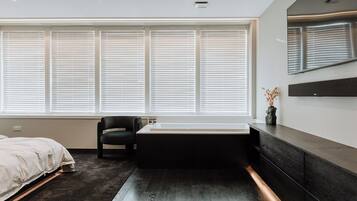 Un dormitorio moderno con un ventanal grande, una silla negra, una bañera y un suelo de madera oscura.