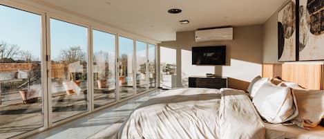 Un dormitorio con un amplio ventanal corredera, una cama con ropa blanca, un televisor de pantalla plana y una obra de arte en la pared.