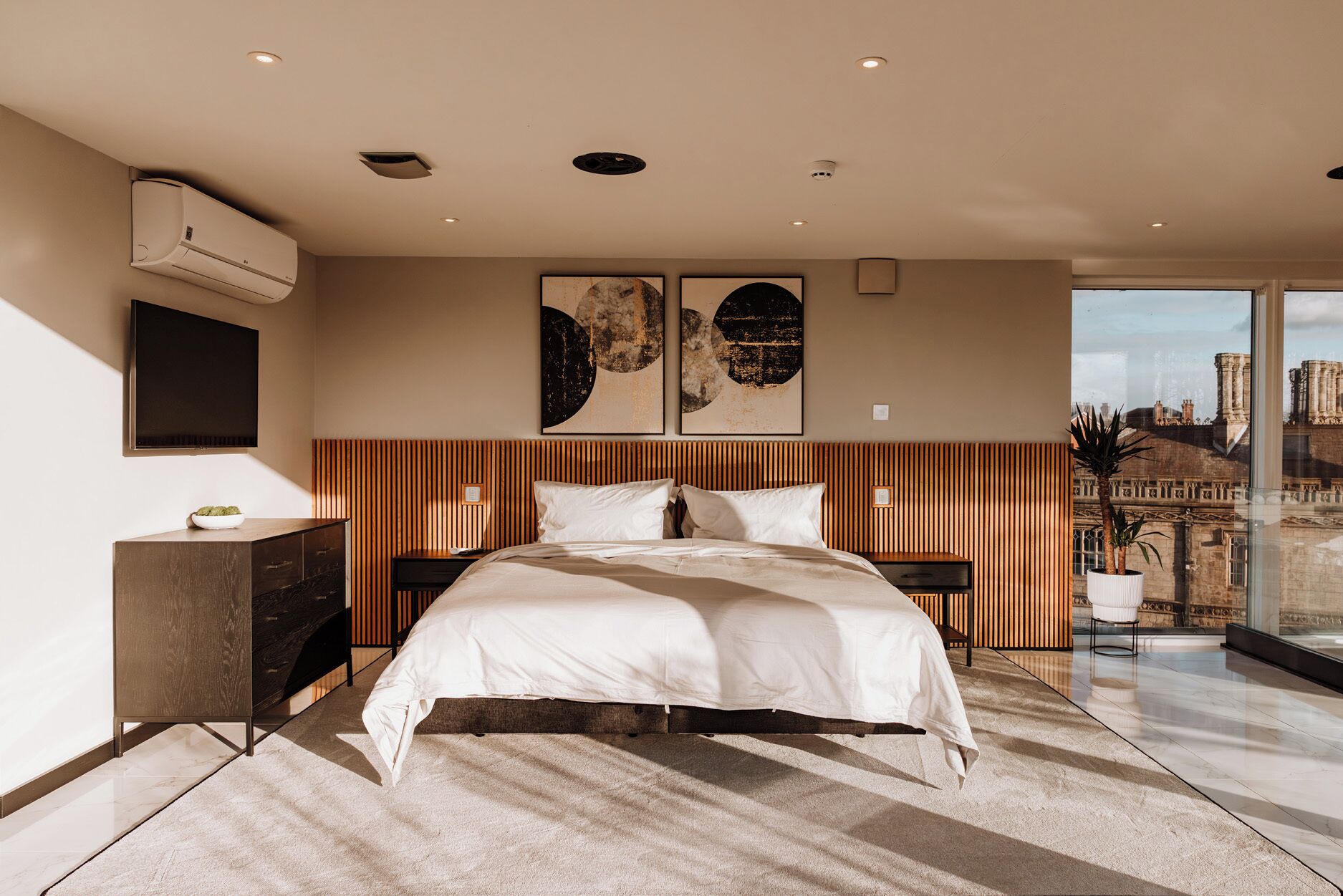 A modern bedroom with a large bed, a flat-screen TV, and a city view.