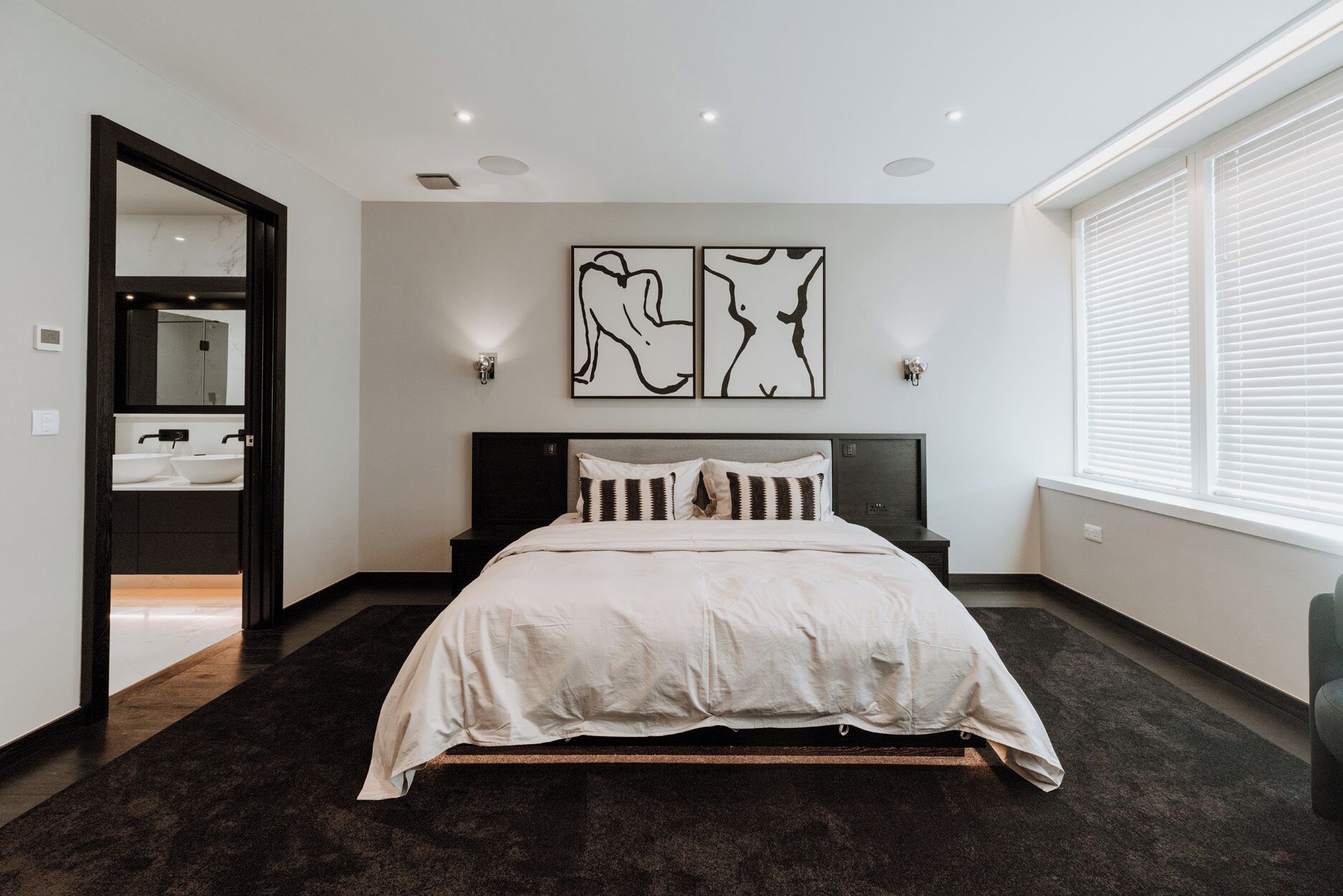 A modern bedroom with a large bed, a dark floor, a wall-mounted artwork, and a window with blinds.