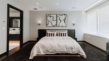 A modern bedroom with a large bed, a dark floor, a wall-mounted artwork, and a window with blinds.