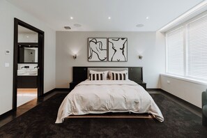 A modern bedroom with a large bed, a dark floor, a wall-mounted artwork, and a window with blinds. - Luxury two Story Penthouse With Roof top Terrace (Shrewsbury)
