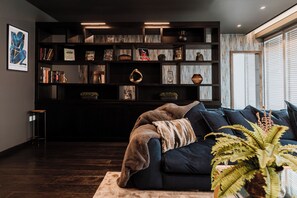 A modern living room with a dark-colored sofa, a bookshelf filled with books and decorative items, and a large window with blinds. - Luxury two Story Penthouse With Roof top Terrace (Shrewsbury)