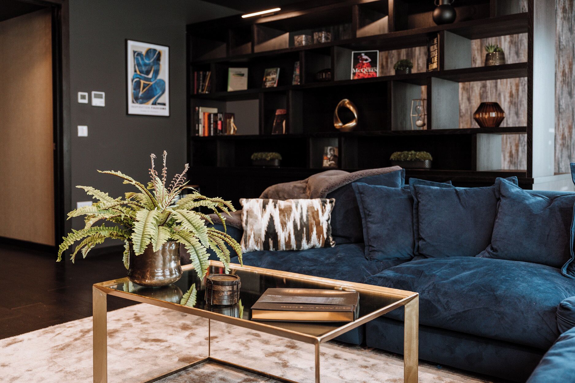 A modern living room with a dark blue sofa, a glass coffee table, and a bookshelf filled with books and decorative items.