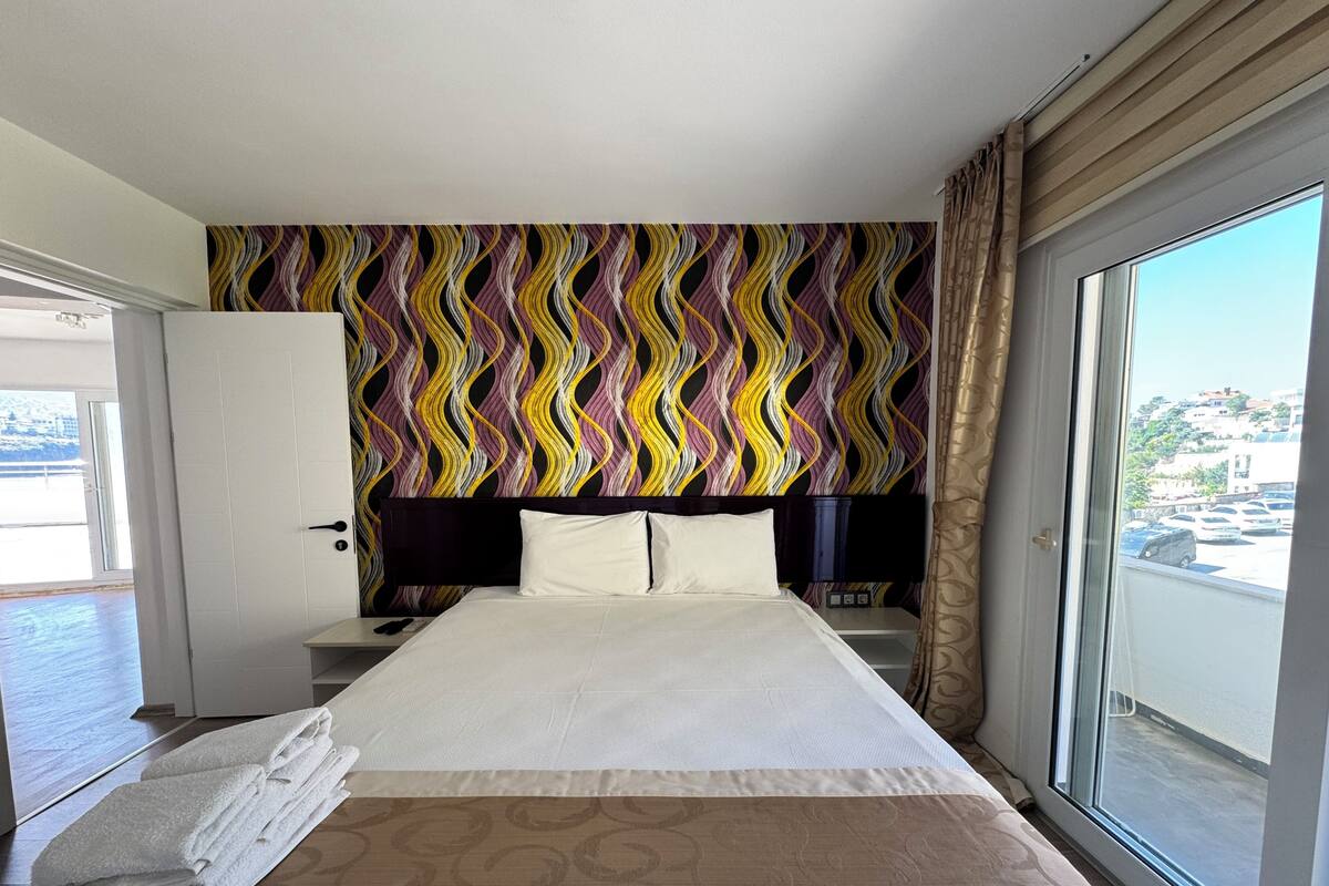 Habitación de hotel con cama, mesita de noche, cómoda y vistas a la ciudad a través de un ventanal grande.
