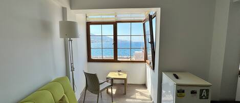 Une pièce avec un canapé vert, une petite table, une chaise, une lampe, un réfrigérateur et une fenêtre donnant sur la mer.