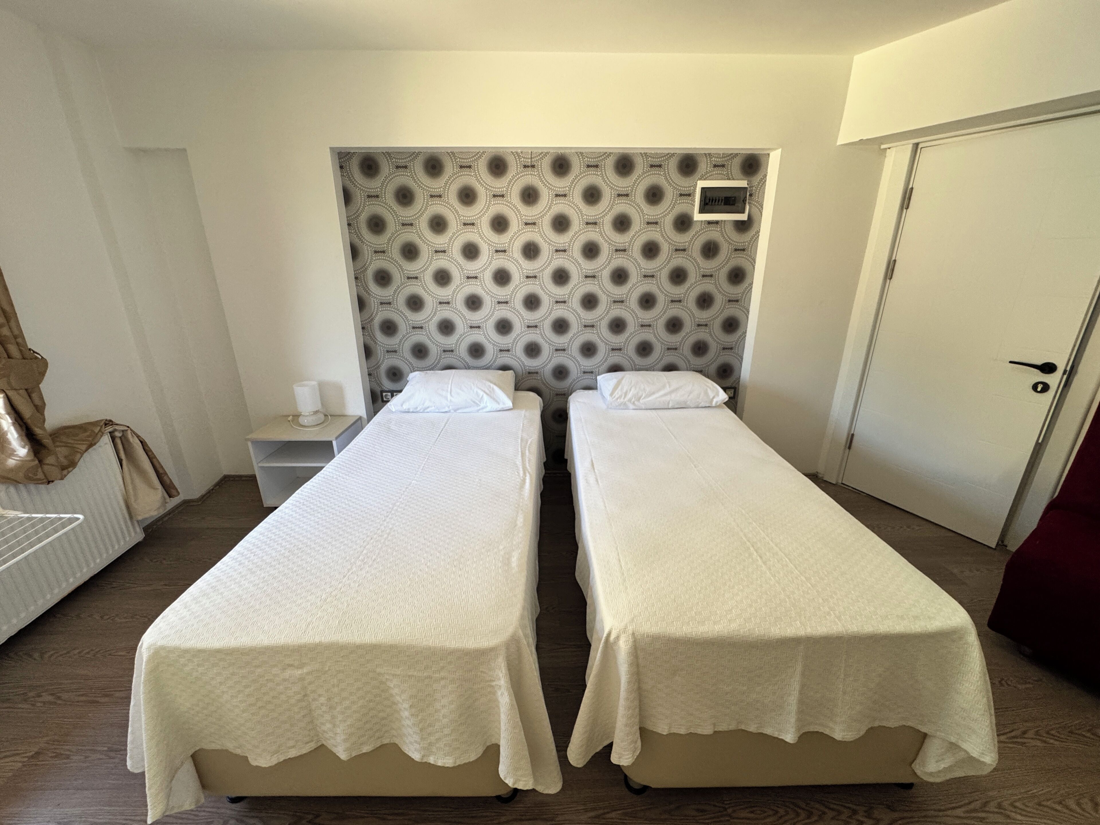 Ein Doppelzimmer mit zwei Einzelbetten, einem gemusterten Kopfteil, einem Nachttisch mit Lampe und einer Tür.