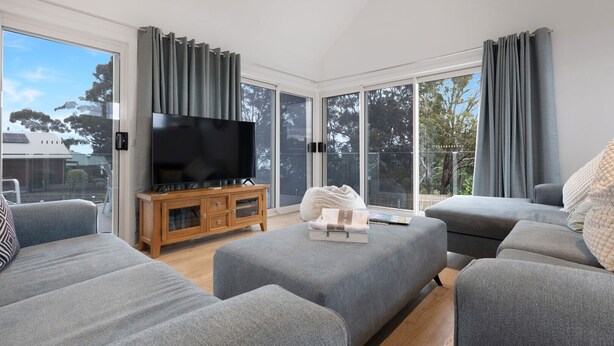 A modern living room with grey sofas, a wooden TV stand, and a large flat-screen TV. - Leighton Bay Views offers a comfortable and stylish base for your stay (Metung)