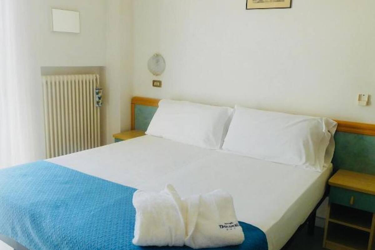 A hotel room with a bed, a nightstand, a radiator, a wall clock, and a framed picture.