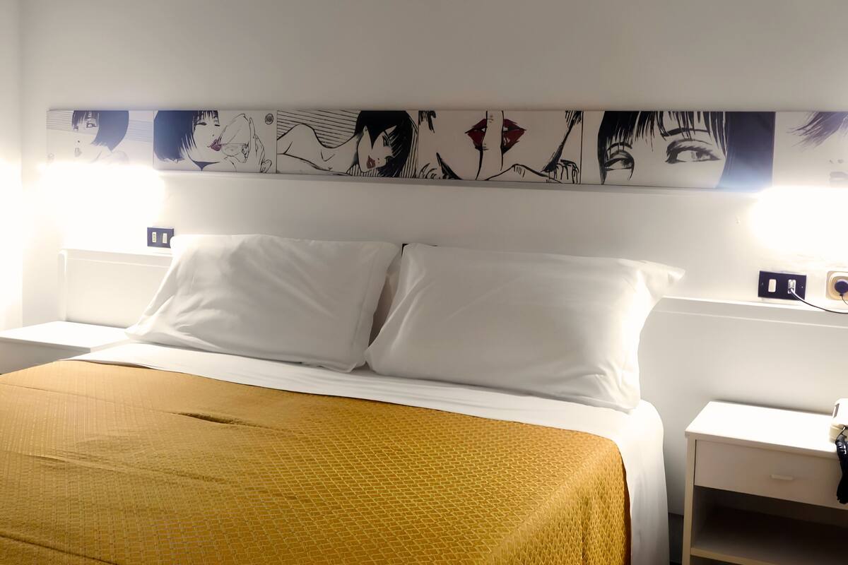 A hotel room with a bed, white pillows, a mustard-colored blanket, a wall-mounted artwork, and a nightstand with a lamp.