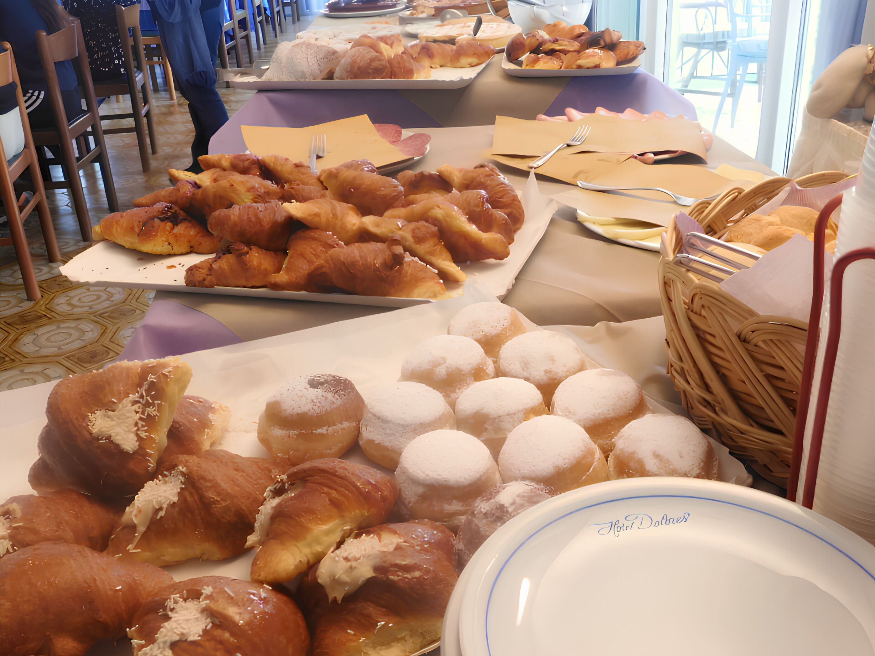 Una mesa de bufé con diversos pasteles y pan, incluyendo croissants y donas, con utensilios para servir y servilletas.