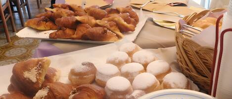 Una mesa de bufé con diversos pasteles y pan, incluyendo croissants y donas, con utensilios para servir y servilletas.