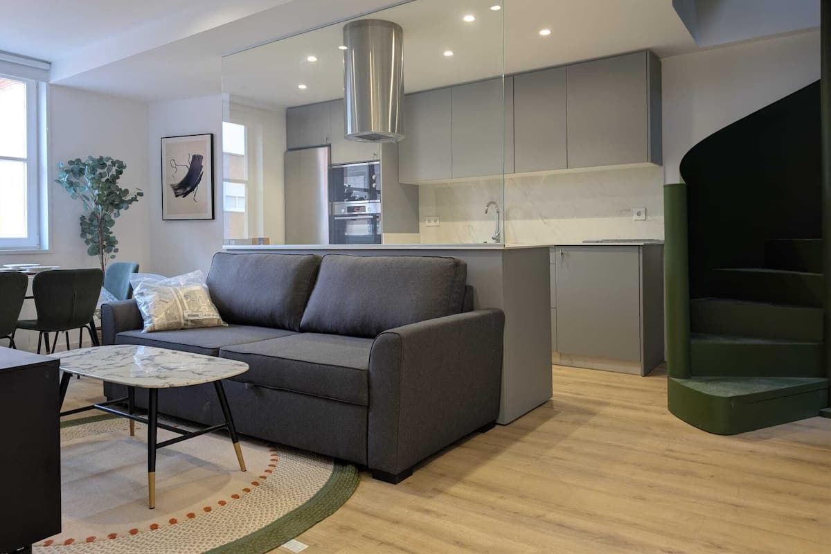 Un salon moderne avec un canapé gris, une table basse ronde et une petite table à manger. On aperçoit, au fond, une cuisine avec des meubles gris et des appareils électroménagers en acier inoxydable.